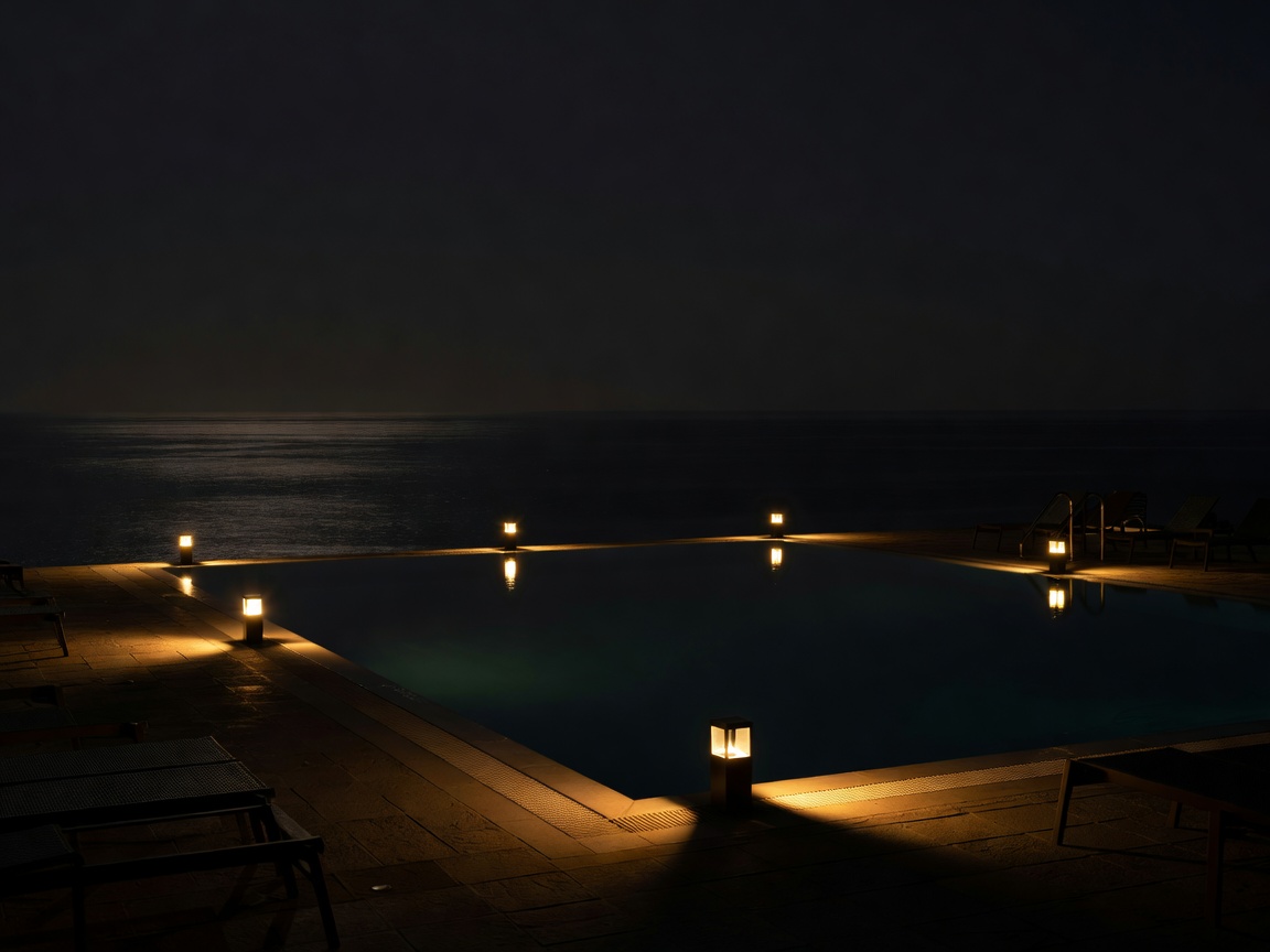 Hotel pool terrace at night with low lamps, deep shadows and the sea beyond